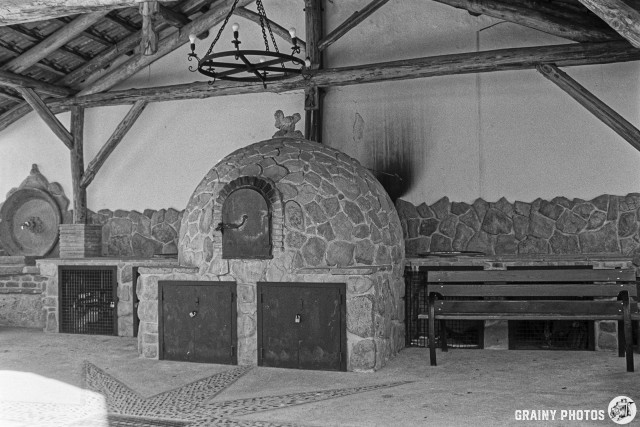 An indoor space featuring a comunal stone bread oven with a domed top, set against a rustic wooden structure. The walls are adorned with stone, and there’s a bench for seating. A chandelier hangs from the ceiling, adding to the charm of the setting.