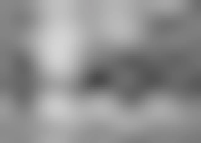 A chapel sits on top of a snow covered hill in a cemetery. Headstones of different shapes and sizes can be seen as well as various large trees. This is a black and white image. 