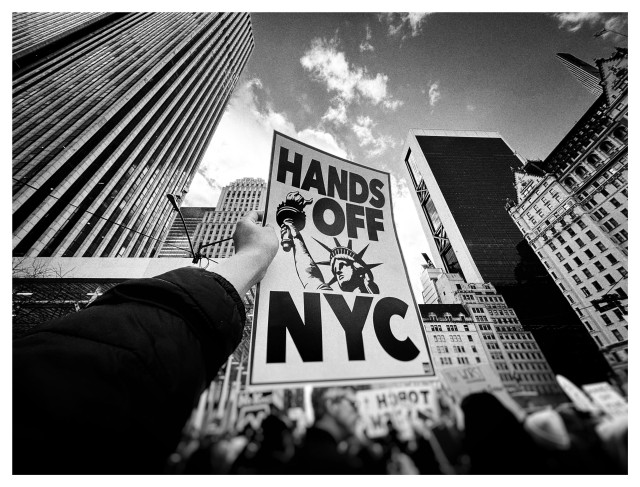 Black and white wide-angle photo of a crowd of protesters dwarfed by high-rise buildings under a partly cloudy sky. The camera is tilted upward so that the buildings are distorted and recede towards upper frame. A protester’s arm in the foreground holds up a sign that occupies the center of the picture and reads “HANDS OFF NYC” with an illustration of the Statue of Liberty. Taken at the NO WAR, NO KINGS, NO ICE rally in New York, Jan 11, 2026.
