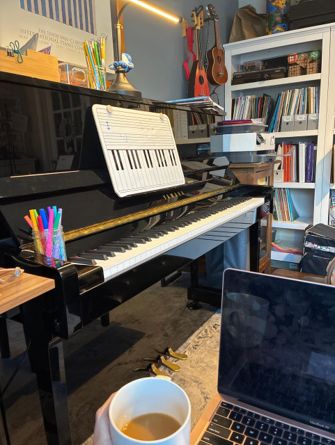A black upright piano in a room with bookcases. A lap holds a computer and a hand holds a cup of coffee.