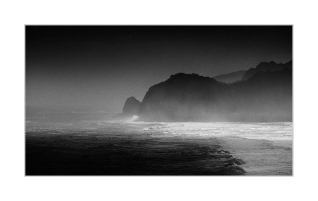 Wide horizontal black-and-white photograph. The scene shows an ocean with layered waves and foam moving diagonally across the foreground, transitioning from darker water on the left to brighter surf on the right. In the midground, sea spray and mist form a low haze along the shoreline. A large, dark headland rises from the water near the center-right, with additional ridgelines receding into softer, lighter tones behind it, while the upper portion of the image is dominated by a dark, evenly graded sky.