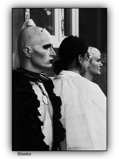 A black-and-white street photograph captures the profiles of three individuals with a distinct queer aesthetic. In the foreground, a person with a shaved head adorned with small decorative spikes and a long, fair ponytail stands out. They wear bold, graphic eye makeup and dark lipstick, dressed in a black ruffled shirt paired with a spiked leather collar and cascading metal chains across the chest. Next to them stands a person in a light, airy tulle outfit, while the composition is completed by a third individual with short, light hair and shimmering glitter on their cheeks. The high-contrast image conveys a sense of pride and uncompromising self-expression, capturing the vibrant atmosphere of the march.