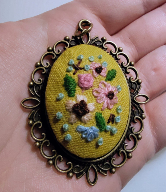 A small embroidery of flowers done on a yellow fabric, set in a bronze pendant, all lying in the palm of my hand. 