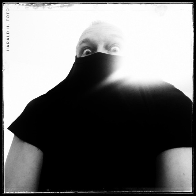 A black-and-white self-portrait of a man who has only pulled his black T-shirt halfway over his head. The image has high contrast, and to the right of the head, a light source is overexposing parts of the image.