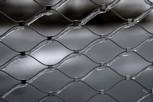 Das Foto zeigt ein vereistes Maschennetz aus Stahl. Es wirkt sowohl robust und industriell als auch ästhetisch und beinahe künstlerisch. Das Eis verleiht dem Stahldraht eine glänzende, kristalline Oberfläche, die das Licht auf interessante Weise reflektiert. So entsteht ein faszinierender Kontrast zwischen der Härte des Metalls und der zerbrechlichen Schönheit des Eises.
 ----- 
The photo shows an icy steel mesh net. It appears both robust and industrial as well as aesthetic and almost artistic. The ice gives the steel wire a shiny, crystalline surface that reflects light in an interesting way. This creates a fascinating contrast between the hardness of the metal and the fragile beauty of the ice.