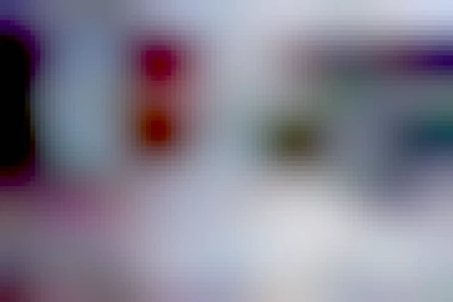 A snowy outdoor scene features a Japanese flag in the foreground, showing a red circle on a white background. In the background, there is a dark-colored building with multiple windows, snow on its roof, and a sign displaying "ART GALLERIES". Two individuals dressed in winter clothing are walking on the snow-covered ground near the building.  