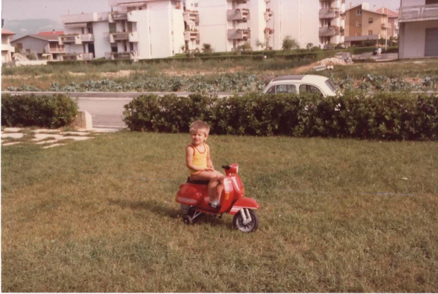A young child (me) sits on a red toy Vespa on a grassy lawn, wearing a sleeveless top and sandals. In the background there are low apartment buildings, hedges, a road with a parked car, and open fields, giving a suburban Italian feel typical of the early 1980s.
