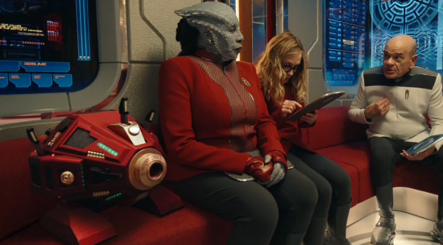 Screen capture from Season 1 Episode 2 of Star Trek: Starfleet Academy, where inside a turboblift, The Doctor, Captain Ake and Commander Lura Thok have a conversation next to Almond Basket, an Exocomp 

The Exocomps are sentient robots last seen as a species in Lower Decks but originally from Star Trek: The Next Generation.  

This exocomp is Maroonish red with gold and black trim and glowing green accents and LEDs.