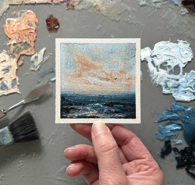 Photo of a hand holding an original seascape oil painting by Tisha Mark, "Soft Peaceful Sunset No. 2601" 3"x3" oil on Arches oil paper (2026). A textured seascape painting with a light blue sky with orange sunset clouds over a dark blue sea with a rocky shore. Color from the sky reflects in the water below. A paint palette with puddles of oil paint, a paintbrush, and a palette knife are visible in the background.
