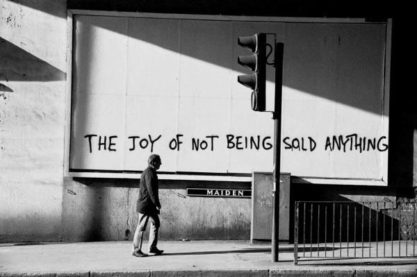 A blank billboard sign in black and white, with graffiti on it "THE JOY OF NOT BEING SOLD ANYTHING"

Underneath it is a partially obscured station sign "MAIDEN" suggesting it may have been taken in Maidenhead, UK

A man walks underneath the billboard, unknowing he is being photographed