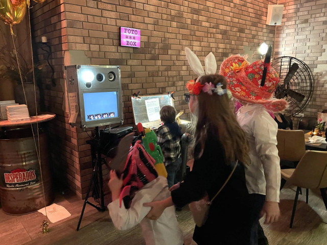 Menschen in Verkleidung stehen vor einer Fotobox. Sie haben sich mit allerhand albernen Hüten und Hasenöhrchen dekoriert und warten drauf das das Gerät ein Foto macht. 