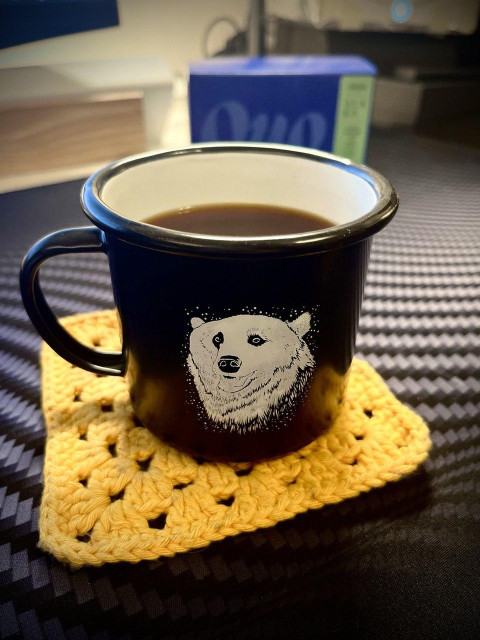 ENG: Photo of an enamel cup with black coffee in it, sitting on a yellow crocheted coaster. a coffee package from the roaster Quo can be seen blurred in the background.

FI: Kuva enamelimukista, jossa on mustaa kahvia. Kuppi on asetettu keltaisen virkatun kupinalusen päälle. Taustalla näkyy sumennettuna Quo-kahvipaahtimon kahvipaketti.