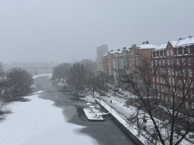 En grå och dimmig bild från St Eriksbron i Sthlm över kanalen. Is med snö mitt i kanalen, öppet vatten löngs strandkanten. Bryggor, gångväg och Hyreshus på höger sida längs med vattnet.