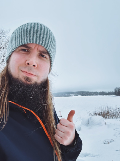 A man with long hair and a beard giving a thumbs-up outdoors in a snowy landscape during winter.