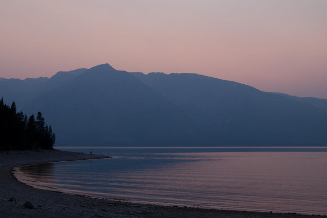 Smoke haze sunset in the Tetons. A shoreline of a lake borders the bottom of the photo and extends to the left side and terminates by a stand of evergreens. Moutmntains in the peach colored haze varely show their divisions across the lake. The sky is cloudless. Colors are muted due to the haze.