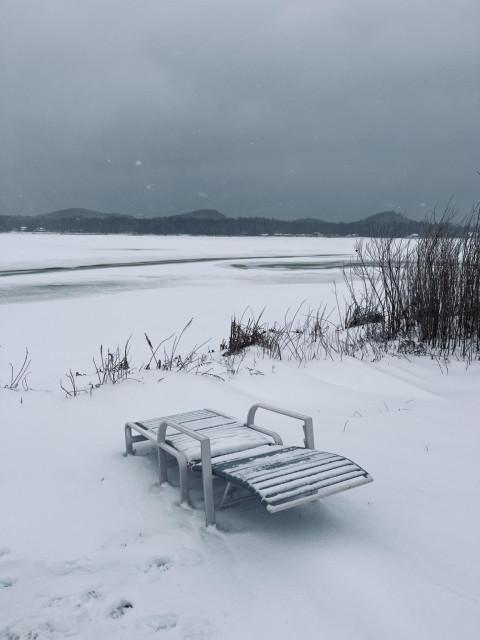 A summer poolside lounge recliner sits at the side of a frozen lake covered in snow during a snowstorm with heavy low -hanging grey clouds.
