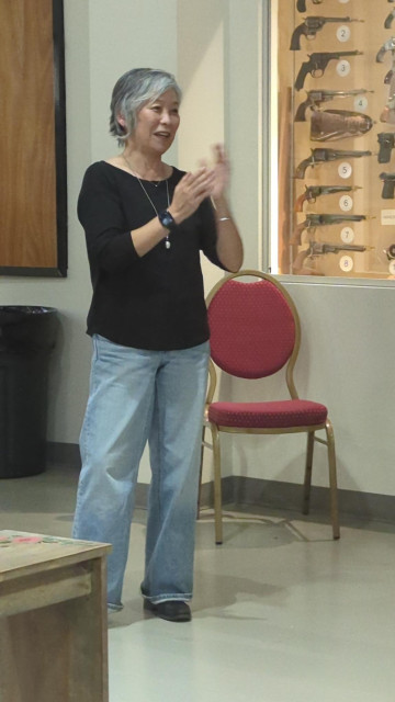 A woman (me) with gray hair in a black sweater is giving a speech. 