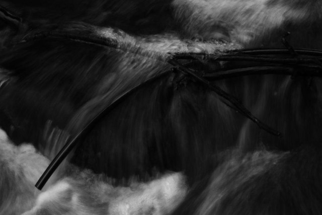 An image of a small waterfall with branches laying as a cross over it. It makes the water slow down a bit to flush over and some bubbles was made due to the branches. The somewhat slow shutter speed makes the water look a bit like it has been painted.
