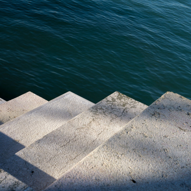 Photo looking edge on at a flight of stone stairs heading down to the left, a sawtooth of shadow-kissed white across the middle of the image below rippling seagreen water.