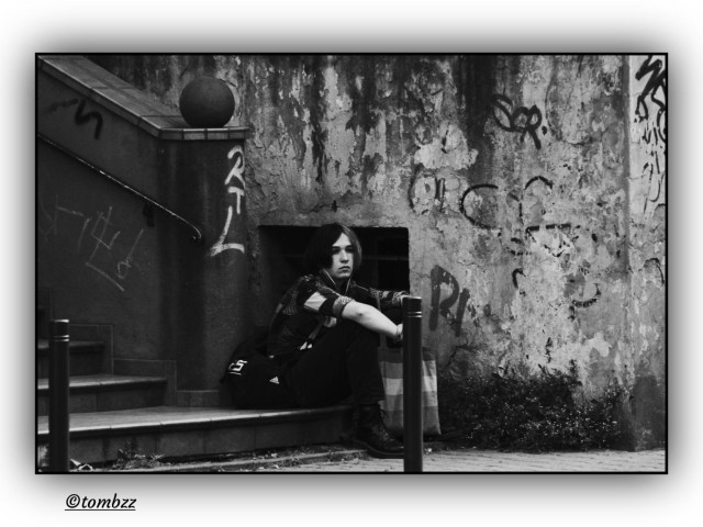 Black and white street photo. A person sits on a step by a worn, graffiti covered wall with layers of tags, letters and random marks. Shoulder length hair split into two colors, one side light, the other dark. They’re wearing dark pants, heavy boots and a T shirt that looks like it would be colorful in real life. In one hand they’re holding a fabric tote in rainbow colors. Next to them a basement window with bars, a bit of the stairs with a round railing post. The body slightly leaning forward, hands resting on the knees, a moment of pause, maybe reflection. The whole scene feels raw, but quietly affirming.
