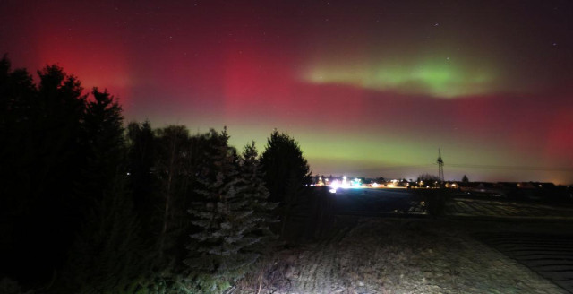 nächtlicher Blick über Wiesen und Bäume zu einem Nachbardorf, der Himmel darüber leuchtet stark in dunkelrot und hellgrün