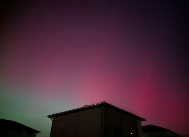 The night sky glows with a soft pink and purple aurora, creating a serene backdrop above a suburban neighbourhood. The silhouette of a house roof is visible in the foreground, adding depth to the scene. Stars twinkle faintly in the sky, the Plough among them, enhancing the tranquil and mystical atmosphere.