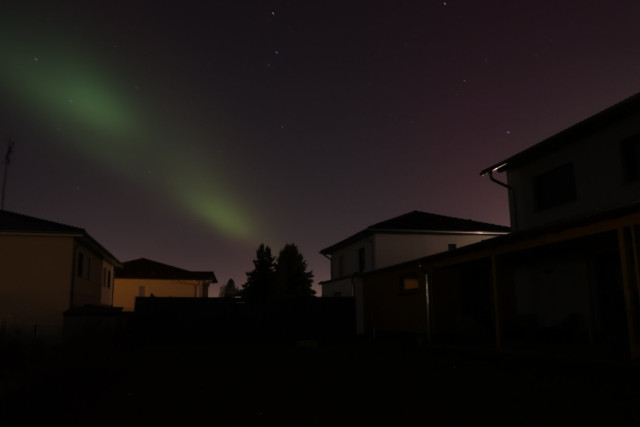 A striking green beam of the Aurora Borealis stretches across the night sky, illuminating the darkness above a residential area. The silhouettes of houses and trees are visible below, providing a sense of scale and location. The vibrant green glow contrasts beautifully with the darker sky, capturing the ethereal beauty of the Northern Lights.