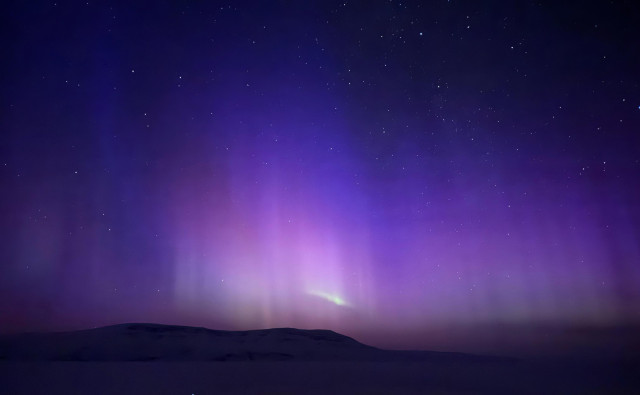 Blue, red, and a tiny bit of green aurora fill a starry sky above the silhouette of hills across the frozen bay. 