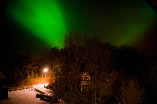 Rechts bis zur Mitte Bäume und ein Haus dahinter. Mitte Links Schnee ein weg eine Dorflampe. Es ist Dunkel. 2 Streifen Nordlichter Vförmig in grün im Himmel.
