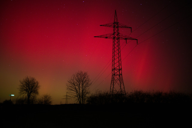 Bäume und Gestrüpp als Silluette unten. Ein Stromast dominant ziemlich mittig. Die Leitungen führen in die Ferne mittig unten. Polarlichter rot in der Ferne leicht grün. Links kaum zu sehen leuchtet ein Wegweiser.