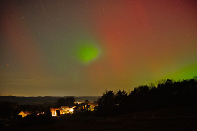 Links unten leuchtet ein Dorf. Rechts Baum Silhouetten mit leichter Steigung zum Rand. Rot grüne Polarlichter. Rote Streifen. Ein grüner Fleck in der Mitte.