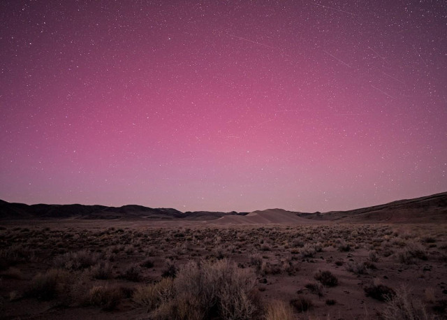 A color landscape photo of a red aurora over a sagebrush plain. In the distance is a large sand dune, but it looks small because of the wide-angle lens. There's lots of stars in the sky and lots of streaks of satellites reflection sunlight shortly after dusk.