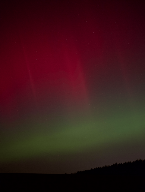 Red/violet smudges in the sky underlined by a stain of green just above a horizon in pure black. Stars shine through in the sky.