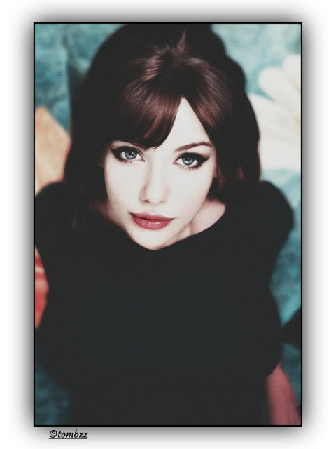 Portrait of a woman with fair skin, photographed on film in soft, slightly surreal light. She wears a simple dark top without embellishments that keeps the focus on her face. Her hairstyle is intricate: a short, sixties‑inspired front and a pinned‑up back, all carefully arranged with a clear sense of style. Her makeup is bold, also echoing the sixties in a modern way, with strongly defined eyes and a winged or cat‑eye contour that gives her gaze its intensity. Her lips are vivid in color, drawing attention and balancing the composition of her face.

Her green eyes look straight ahead, intense but not harsh. There is something more in that gaze, a veiled sensuality, maybe a hint of melancholy. Something that leaves room for personal interpretation.

The background is an abstract composition with floral elements, full of color that contrasts with her dark clothing. It doesn’t distract but surrounds her gently, as if she were part of a larger story that can’t be fully decoded.