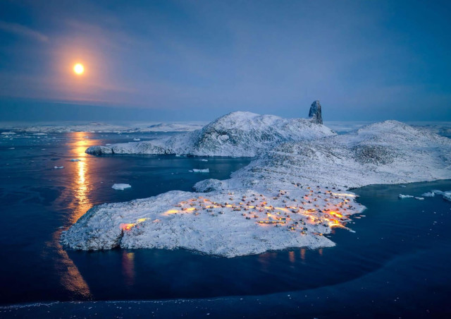 A stunning winter landscape features a moonlit night over snow-covered hills and a coastal village in Greenland illuminated by warm lights.