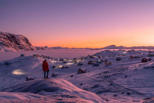 A person in a red jacket stands on a snowy hill overlooking a colorful village at sunset, with mountains in the background.