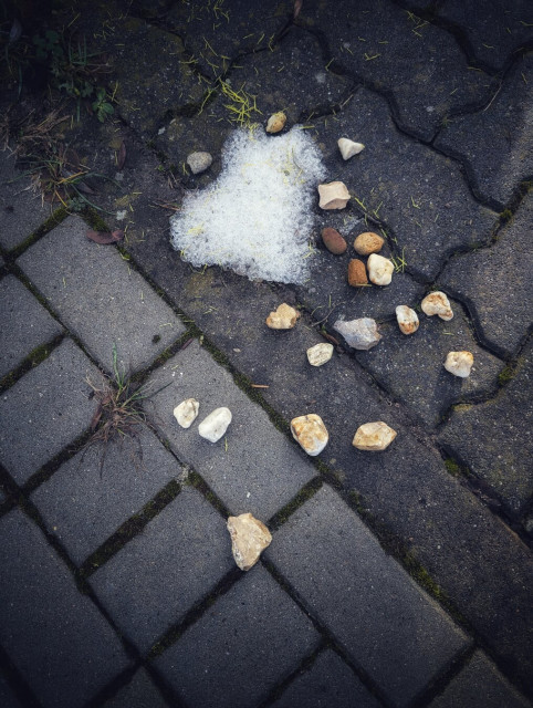 A close-up, overhead view of a paved pathway made of rectangular grey bricks. In the centre of the image, there is a small, heart-shaped patch of snow surrounded by a scattering of multi-coloured stones. The stones vary in size, shape, and colour, ranging from light beige to darker brown. Some small patches of grass and moss are visible growing between the bricks, adding a touch of green to the otherwise grey and earthy tones of the scene.