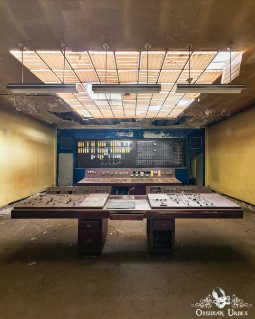 A dusty, abandoned control room with an old control panel and wall of switches beneath a partially damaged, skylit ceiling.