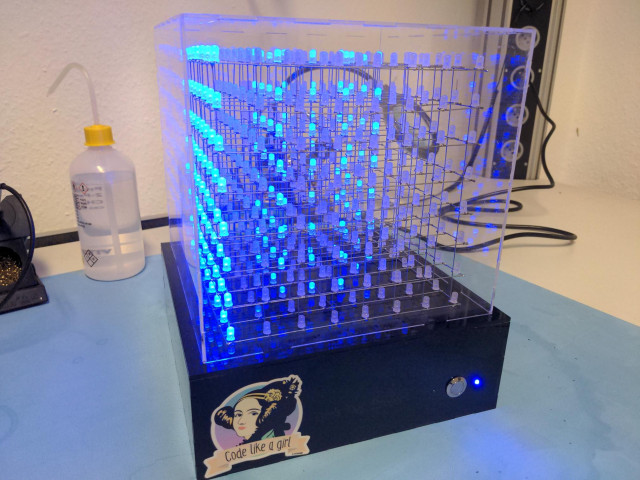 an 8x8x8 LED cube with blue lights. it has a black electronics compartment with a sticker "code like a girl" and engravings "girls day 2020" on the front. there is also a button to switch animation modes.