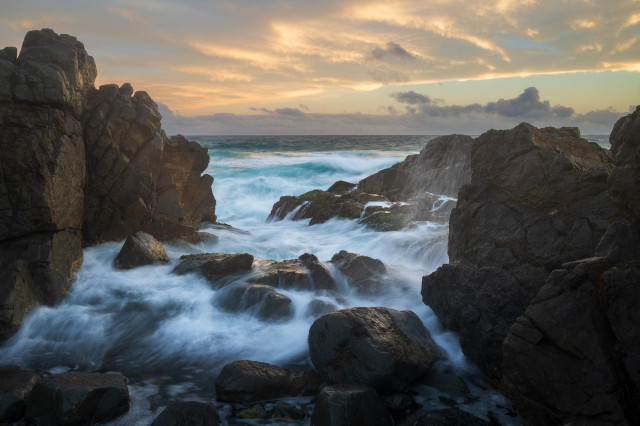 A rugged coastal scene at golden hour. Dark, weathered rock formations frame the image on both sides, forming a natural corridor toward the open sea. Long-exposure water flows silkily around the rocks, turning the waves into soft, milky streams that contrast with the sharp, textured stone. The ocean beyond is restless but controlled, while the sky glows with warm pastel tones - peach, gold, and pale blue, layered with gentle clouds, suggesting either sunrise or sunset. The overall balance is between power and calm, permanence and motion.