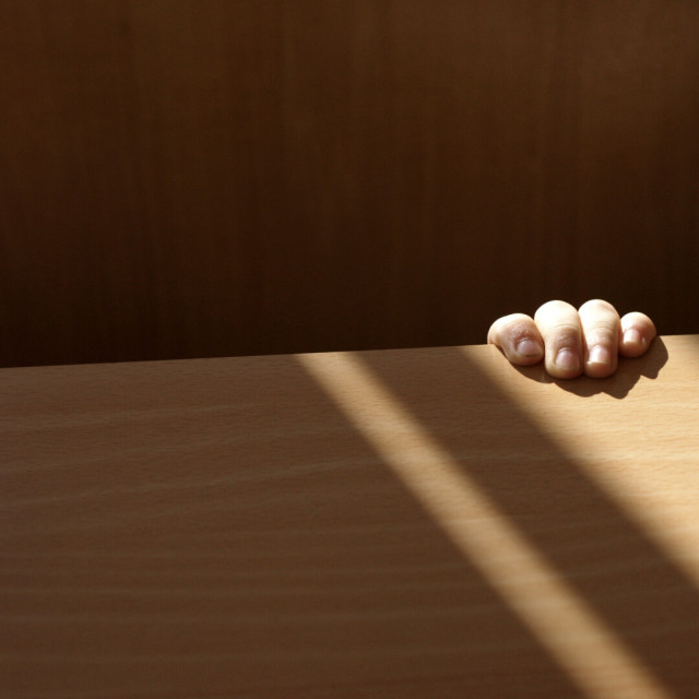 A photo divided in two: a section of wooden chair at the top and part of a lighter wooden table at the bottom. At the right four small fingers grip the tabletop, bathed in a strip of sunlight.