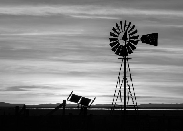 A landscape photo of a solar panel and a windmill silhouetted against a bright sky of horizontally streaked clouds. The solar panel is low in the foreground just right of center. The tall windmill sits on the right. The tail vane of the windmill is pointed toward the right and the large fan toward the left. The sky is streaked in many layers of dark and light clouds. A sliver of a lake is seen cutting from left to right just above the nearly black foreground. The windmill blades and tail vane has many holes in it from irresponsible shooters. 