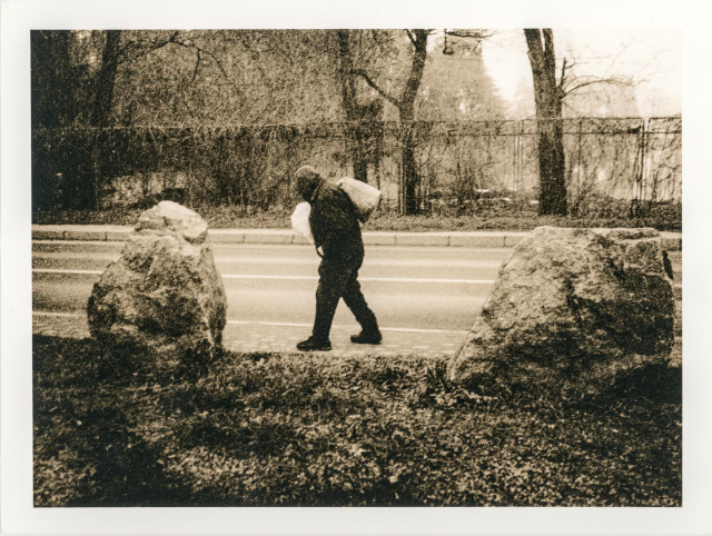 This lith print depicts a solitary figure walking along a roadside, burdened with bags slung over their shoulders. The person is framed between two large stones in the foreground, which give the scene a sense of weight and enclosure. Bare trees and a blurred fence recede into the background, merging with the road in a soft, unstable way.

The characteristic lith printing effect transforms the image into a dense, gritty surface: shadows bloom, highlights appear chalky, and textures dominate over fine detail. The figure seems partially absorbed by the surroundings, as if emerging from or dissolving into the landscape