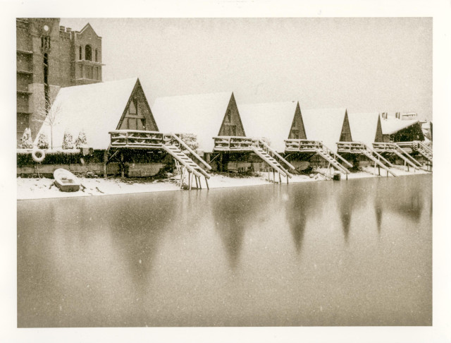 A row of small A-frame cabins lines the frozen edge of a body of water. Their steep roofs and wooden staircases are covered in fresh snow, creating a repeating triangular rhythm across the scene. The water in the foreground is smooth and dark, reflecting the pale silhouettes of the cabins like blurred shadows. Snow falls gently, softening the contrast between architecture and landscape. In the background, a larger brick building looms faintly, adding a quiet sense of scale and distance