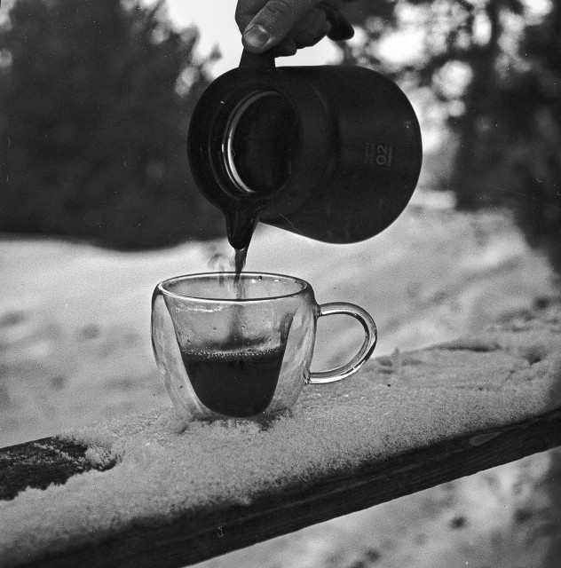A black-and-white photograph shows a hand pouring a hot drink from a small kettle into a transparent glass cup. The cup sits on a snow-covered wooden railing outdoors. Steam rises gently from the liquid, contrasting with the cold, wintry surroundings. The background is softly blurred, with snow and trees forming a quiet, subdued backdrop, emphasizing the intimate moment of warmth against the cold.