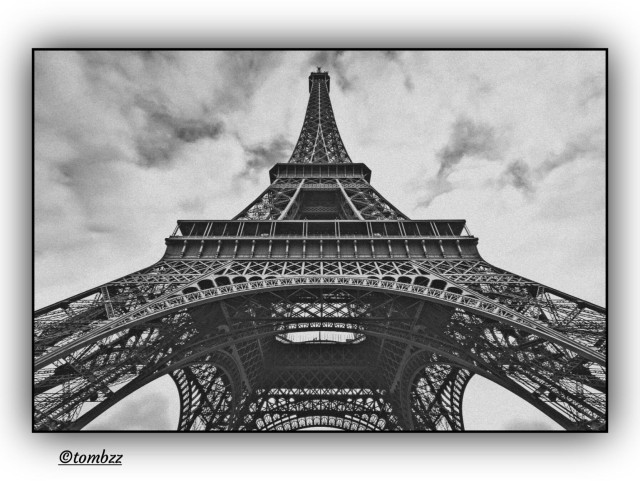 Black and white analog photograph of the Eiffel Tower captured from an exaggerated worm’s-eye perspective. The massive base dominates the frame, pushing outward toward the edges, while the top recedes sharply, losing weight and clarity. The iron lattice forms a dense, almost abstract structure that pulls the eye upward. The background is filled with a dramatic sky and scattered clouds, amplifying the tension between architectural weight and atmospheric lightness. A subtle vignette enhances the sense of focus and depth, giving the image a monumental yet dynamic character, as if the tower were in motion, retreating from the viewer.