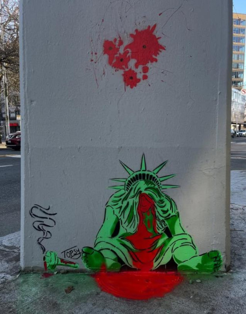 Street art stencil by Topsy in Seattle, USA, showing the Statue of Liberty slumped on the ground beneath a concrete overpass pillar, her head bowed and crown spikes visible, with bright red paint dripping from her face and pooling like blood on the sidewalk; red splatters appear higher on the wall, while the figure is painted in vivid green and black outlines, creating a stark anti-ICE protest image.