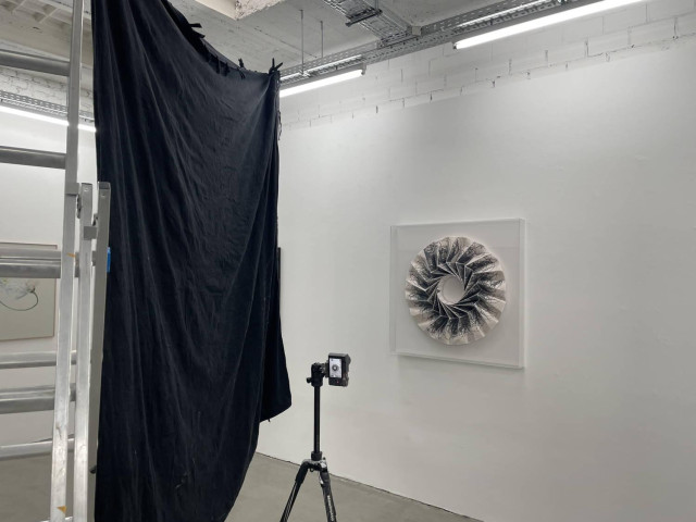 in einem Raum mit grauem Betonboden und weissen Wänden hängt ein geometrisches, rundes Kunstwerk aus bedrucktem Papier, Es ist zu seinem Schutz in einem Plexiglasgehäuse, das mit seinen Spiegelungen schwierig macht, das Kunstwerk ohne Störungen zu fotografieren. Daher ist links von dem Kunstwerk ein Schwarzes Moltontuch aufgehängt, das einen Schatten auf das Plexiglas wirft und so die Spiegelungen unterdrückt