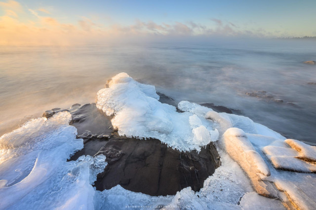 A photos showing sunrise over Lake Superior. In the foreground, a curved slot cut through the ice exposes the black basalt and that contrasts with the ice which is blue in the shadows and yellow on the highlights. Waves wash into the slot from the left and flow down angled basalt to the right. The overall color cast of the image is yellow with blues in the shadows. Sea smoke, which is fog, covers the big lake all the way to the empty horizon. 