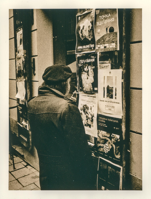 This lith print has a raw, tactile presence, with gritty grain and uneven tones that feel almost alive on the paper. The scene shows an elderly man from behind, standing close to a wall covered with layered posters and announcements. His dark coat and hat merge into the deep shadows, while the lighter paper of the posters flickers with worn textures and fading ink.

The lith process exaggerates contrast and imperfection: blacks bloom softly, highlights break into speckled whites, and the entire surface feels weathered, as if the image itself has aged along with its subject. There is a quiet intimacy in the moment — the man absorbed in reading, smoking, pausing in the flow of the city.